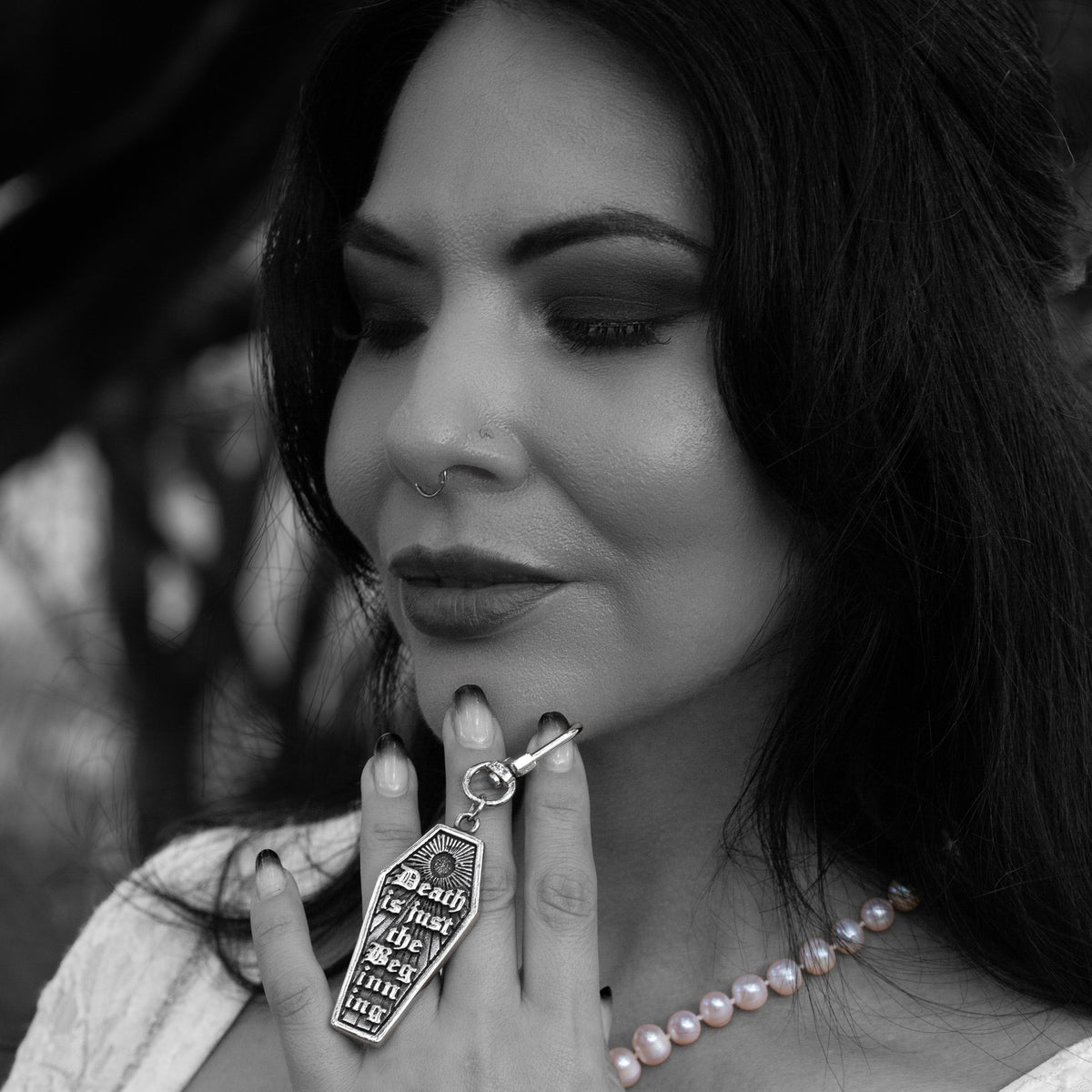 A close-up black and white shot of a woman holding a stainless steel coffin-shaped bag charm with gothic text against her finger, wearing a pearl necklace.