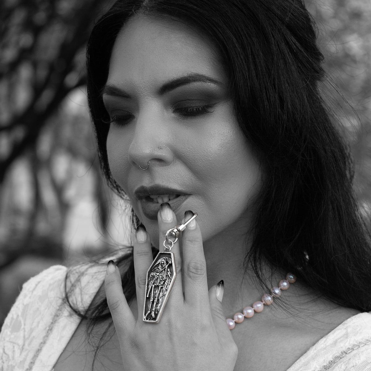 A close-up black and white photo of a woman holding a silver coffin-shaped bag charm keychain with a grim reaper figure, wearing a pearl necklace and dark hair.