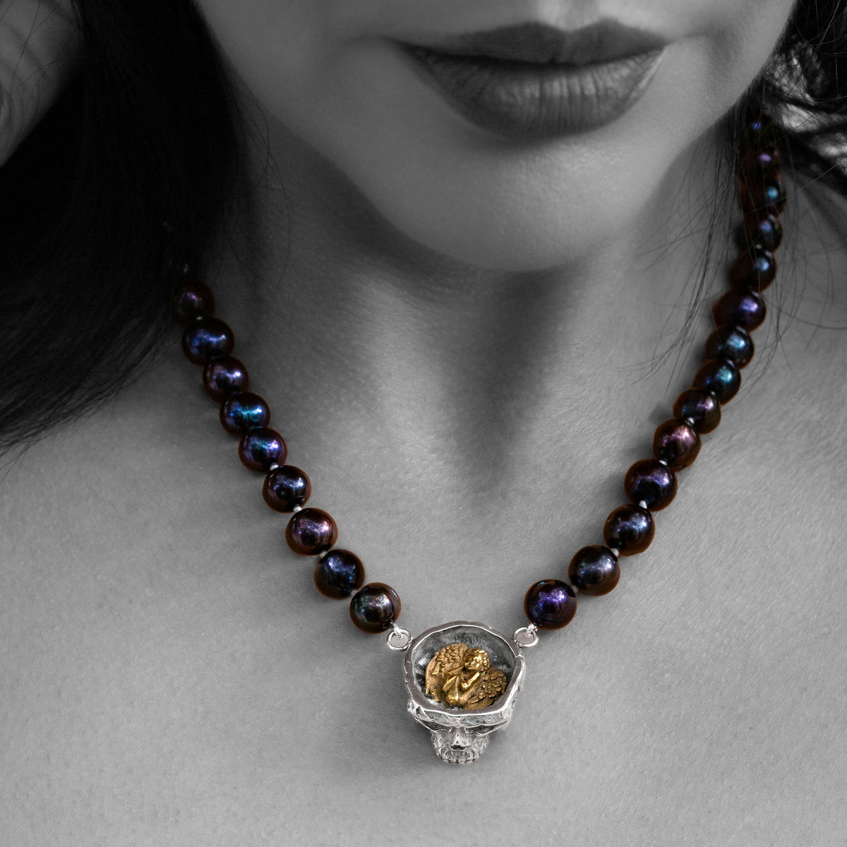 A close-up of a model wearing a necklace with dark iridescent beads and a silver skull pendant containing a gold angel, shown against a black-and-white background with the necklace colours highlighted.