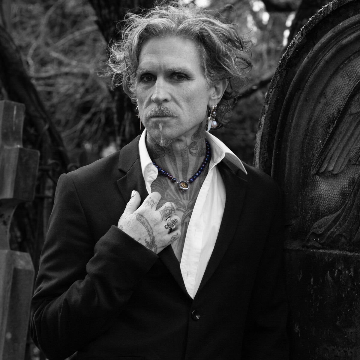 A black and white portrait of a man with wild hair, a goatee, and visible tattoos, wearing a dark suit jacket and open white shirt, standing outdoors near a large stone monument.
