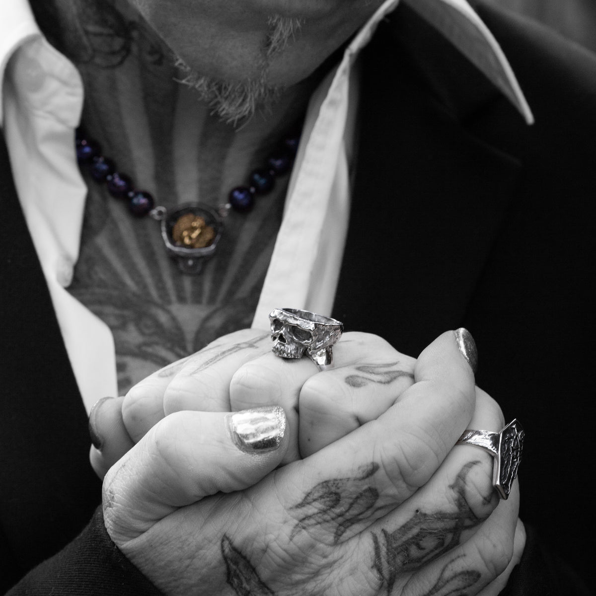 A close-up of a person's tattooed hands clasped together, wearing a chunky silver skull ring and a matching silver ring on the other hand, with a black suit jacket and white shirt in the background.