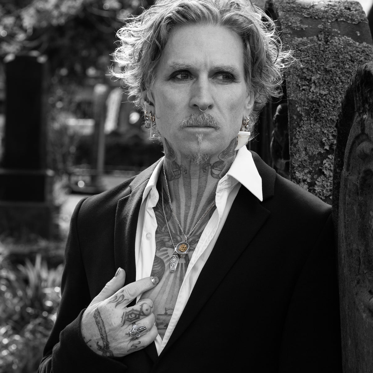 A black and white portrait of a tattooed man with wavy hair, wearing a dark blazer over an open white shirt, leaning against a textured stone wall outdoors.