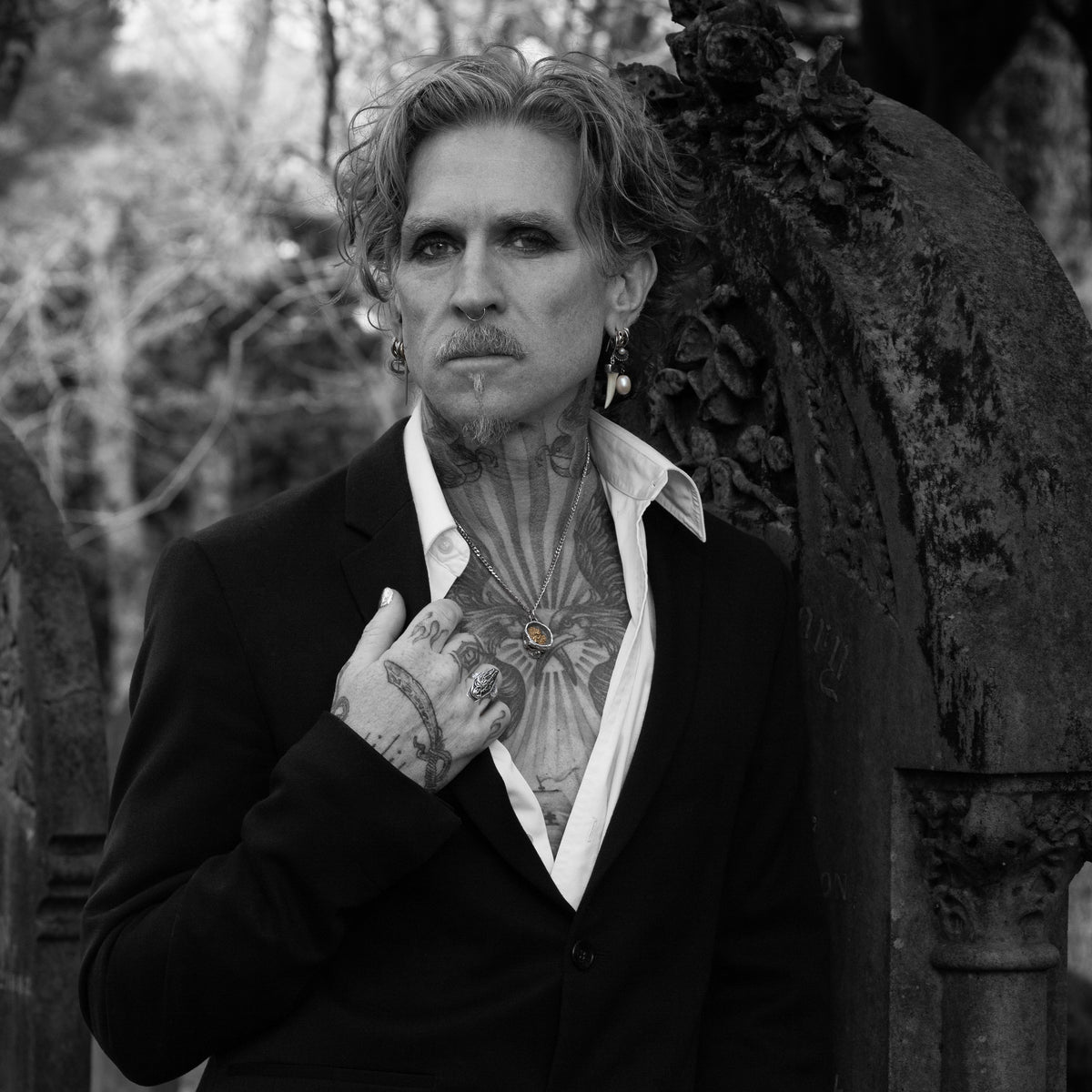 A black and white portrait of a man with tattoos wearing a black suit jacket and open white shirt, standing in front of a weathered stone structure.