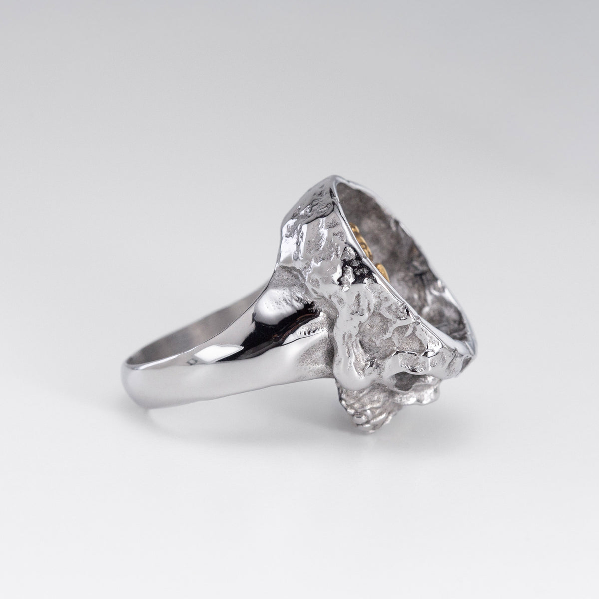 A silver ring featuring a highly textured, organic skull-like top with a polished band, photographed on a clean light gray background.