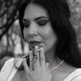 A close-up black and white photo of a woman holding a silver coffin-shaped bag charm keychain with a grim reaper figure, wearing a pearl necklace and dark hair.