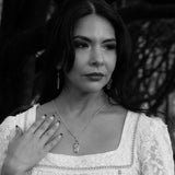 A black and white portrait of a woman wearing a silver gothic coffin shaped pendant necklace and matching drop gothic coffin shaped earrings, with her hand resting on her chest.