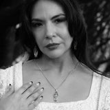 A black and white portrait of a woman wearing a gothic coffin-shaped pendant necklace and matching dangling earrings, with her hand resting near her chest.