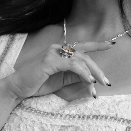 A close-up lifestyle shot of a woman wearing a textured silver ring and a silver necklace with a small orange stone, shown in black and white with selective color on the ring and necklace.