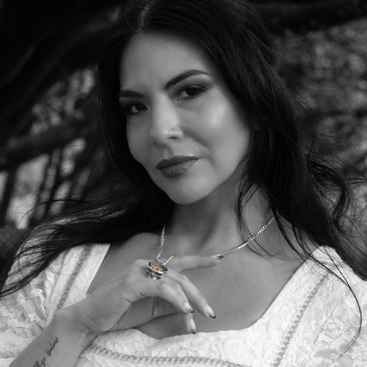 A black and white portrait of a woman wearing a delicate chain necklace with a central skull shaped pendant, resting her hand near the necklace.