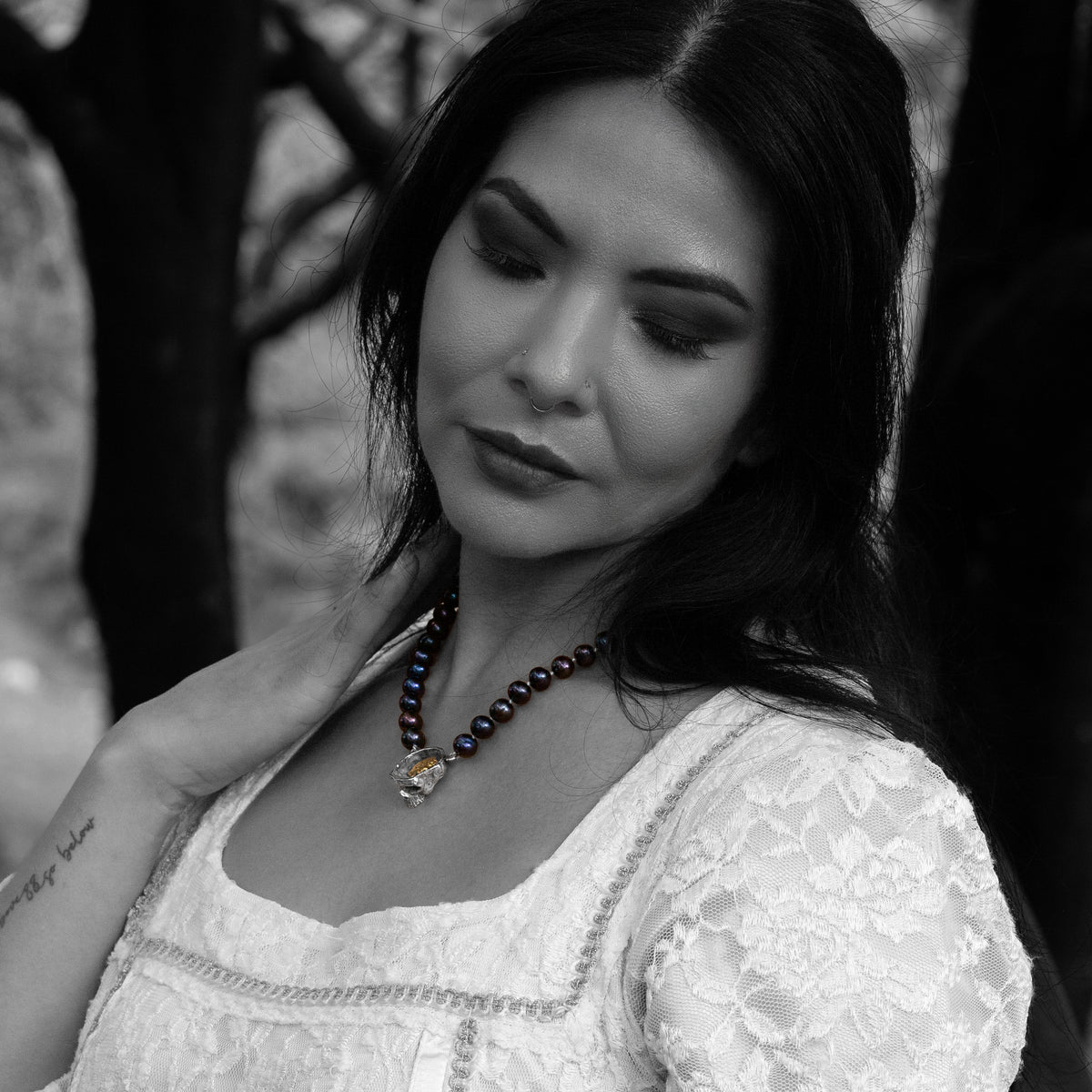 A black and white portrait of a woman wearing a black pearl necklace with a central skull pendant, set against a dark, blurred outdoor background.