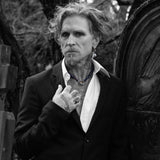 A black and white portrait of a man with wild hair, a goatee, and visible tattoos, wearing a dark suit jacket and open white shirt, standing outdoors near a large stone monument.