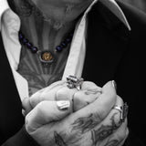 A close-up of a person's tattooed hands clasped together, wearing a chunky silver skull ring and a matching silver ring on the other hand, with a black suit jacket and white shirt in the background.