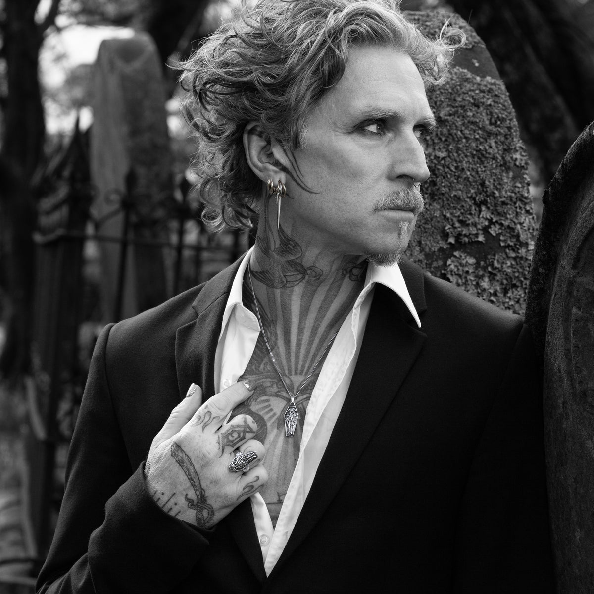 A black and white portrait of a man with curly hair, tattoos, and an earring, wearing a dark suit jacket and open white shirt, posed outdoors against a blurred background.
