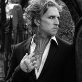 A black and white portrait of a man with curly hair, tattoos, and an earring, wearing a dark suit jacket and open white shirt, posed outdoors against a blurred background.