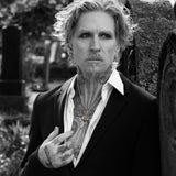 A black and white portrait of a tattooed man with wavy hair, wearing a dark blazer over an open white shirt, leaning against a textured stone wall outdoors.