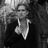 A black and white portrait of a man with tattoos wearing a black suit jacket and open white shirt, standing in front of a weathered stone structure.