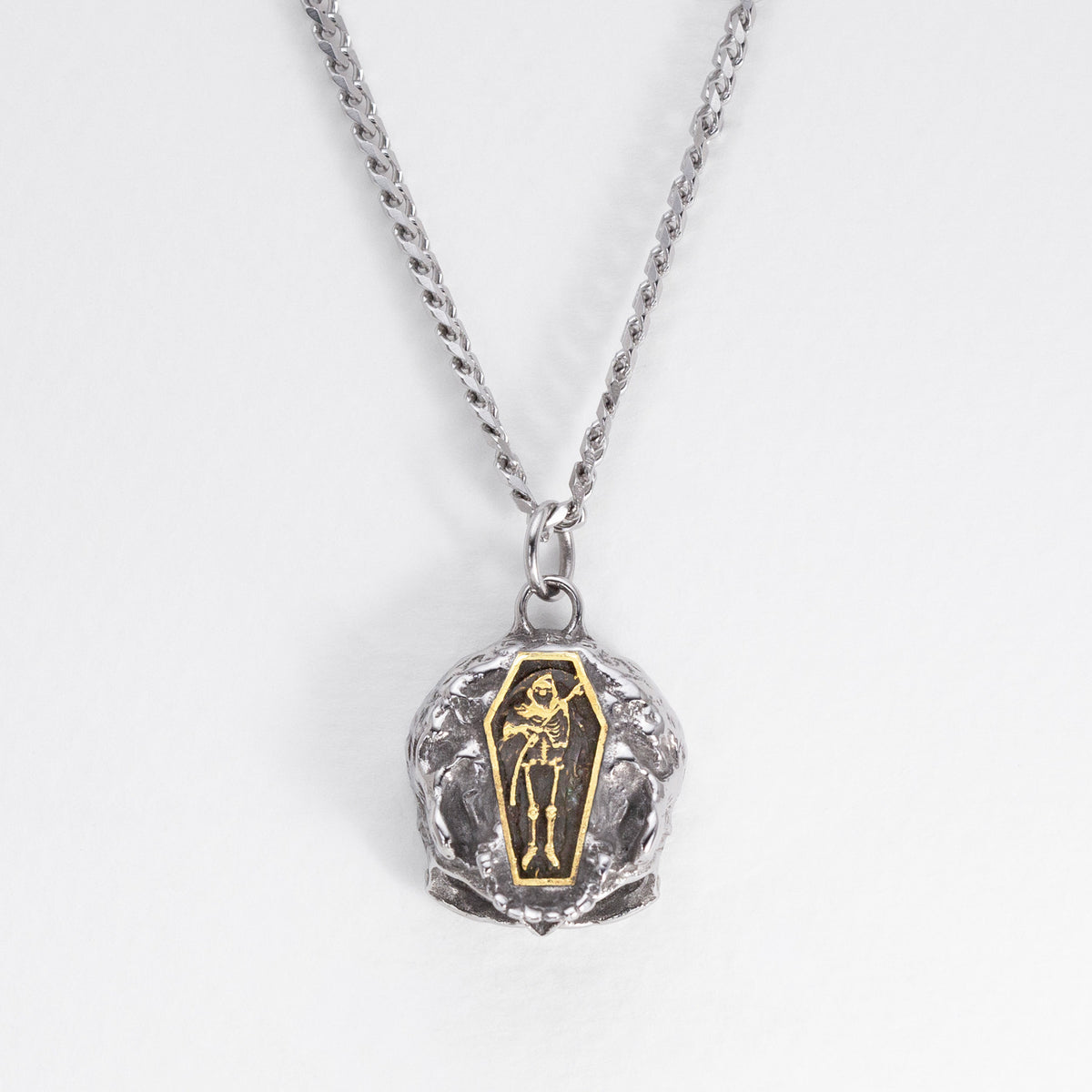 A silver chain necklace with a skull-shaped pendant featuring a gold coffin inlay with a skeleton figure, set against a plain white background.