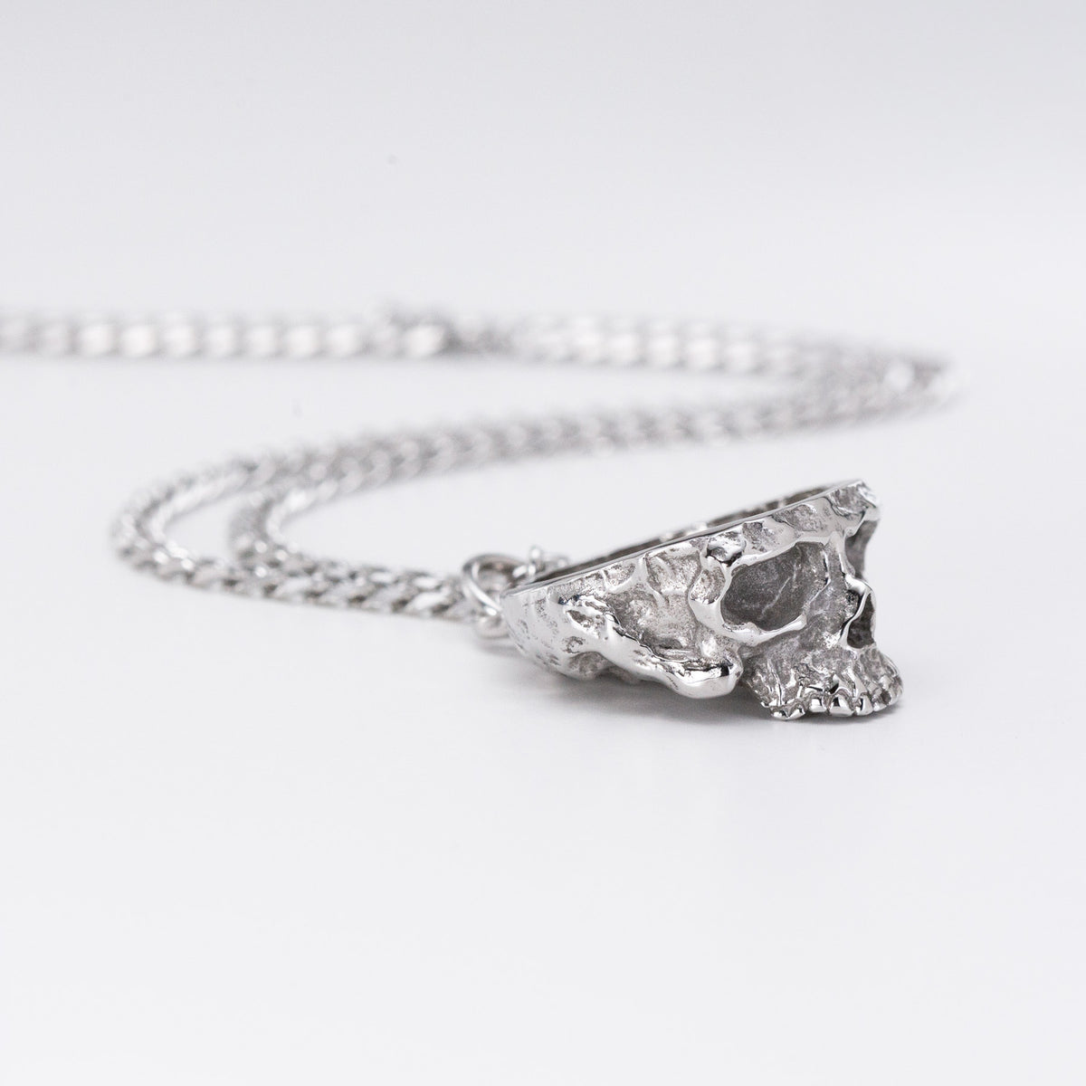 A silver chain necklace featuring a textured, organic-shaped skull pendant, photographed on a clean white background.