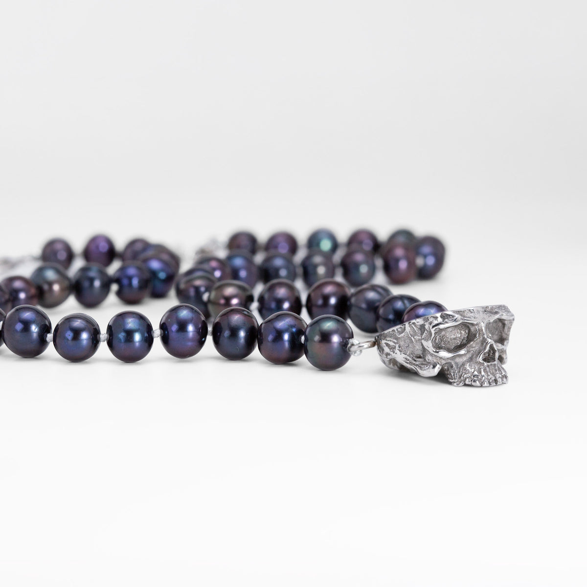 A strand of dark iridescent pearls with a silver skull pendant, photographed on a clean white background.