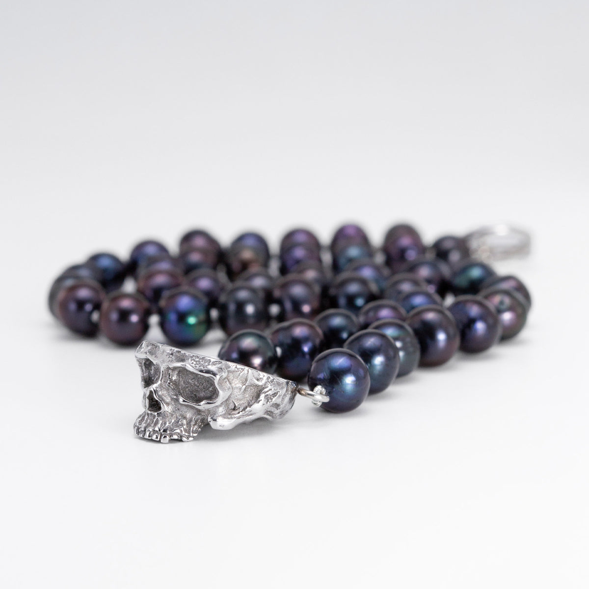 A strand of dark iridescent pearls with a silver skull pendant, photographed on a clean white background.
