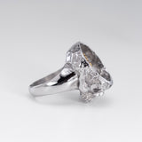 A silver ring featuring a highly textured, organic skull-like top with a polished band, photographed on a clean light gray background.